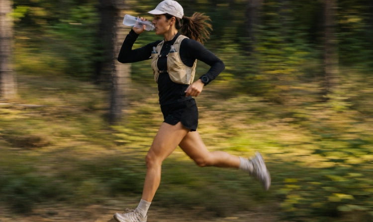 A woman running through a forest while drinking from a water bottle, wearing a beige and black athletic vest and shorts.